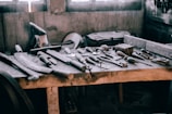 A wooden workbench scattered with various hand tools, including chisels, hammers, and saws. The setting appears rustic and industrial, with a soft light filtering through windows in the background, casting shadows on the tools.