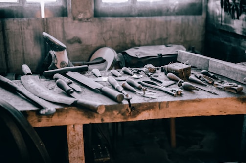 A wooden workbench scattered with various hand tools, including chisels, hammers, and saws. The setting appears rustic and industrial, with a soft light filtering through windows in the background, casting shadows on the tools.