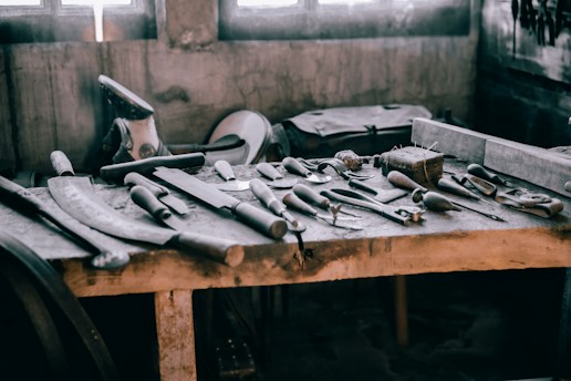 A wooden workbench scattered with various hand tools, including chisels, hammers, and saws. The setting appears rustic and industrial, with a soft light filtering through windows in the background, casting shadows on the tools.