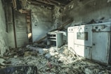 A dilapidated room with significant fire damage. The walls are charred, and the ceiling is partially collapsed, exposing beams. Debris, including papers and wood, is scattered across the floor. A damaged oven and a small desk are visible, covered in soot. The environment conveys a sense of abandonment.