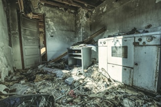A dilapidated room with significant fire damage. The walls are charred, and the ceiling is partially collapsed, exposing beams. Debris, including papers and wood, is scattered across the floor. A damaged oven and a small desk are visible, covered in soot. The environment conveys a sense of abandonment.