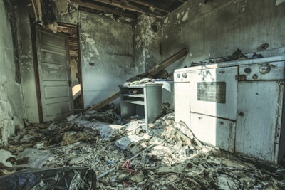 A dilapidated room with significant fire damage. The walls are charred, and the ceiling is partially collapsed, exposing beams. Debris, including papers and wood, is scattered across the floor. A damaged oven and a small desk are visible, covered in soot. The environment conveys a sense of abandonment.