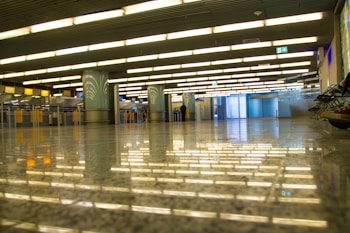 A spacious indoor area with rows of fluorescent lights reflecting on a shiny, polished floor. The area is mostly empty, with a few visible counters and columns. There is minimal foot traffic with a couple of indistinct figures in the distance.
