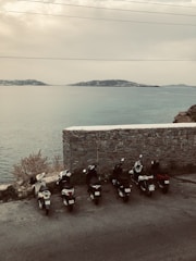 Several scooters are parked in a row on a roadside overlooking a serene body of water. In the background, there is a stone wall and a view of distant hills across the water under a cloudy sky.