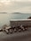 Several scooters are parked in a row on a roadside overlooking a serene body of water. In the background, there is a stone wall and a view of distant hills across the water under a cloudy sky.