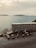 Several scooters are parked in a row on a roadside overlooking a serene body of water. In the background, there is a stone wall and a view of distant hills across the water under a cloudy sky.