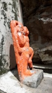 An orange statue of a figure is positioned against a rough stone backdrop. The statue appears weathered with visible chips and wear on its surface. The stone wall behind shows signs of aging with black stains.