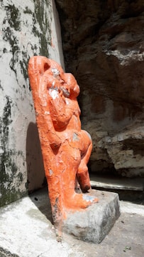 An orange statue of a figure is positioned against a rough stone backdrop. The statue appears weathered with visible chips and wear on its surface. The stone wall behind shows signs of aging with black stains.