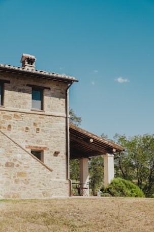 A rustic stone house with a tiled roof stands prominently against a clear blue sky. The structure features two visible windows and a covered patio supported by wooden beams. The surrounding area includes trees and a grassy landscape, giving a serene countryside ambiance.