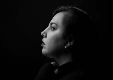 A monochromatic portrait featuring a woman in profile view against a dark background. Light highlights her facial features, creating a dramatic contrast. She wears an earring and dark clothing.