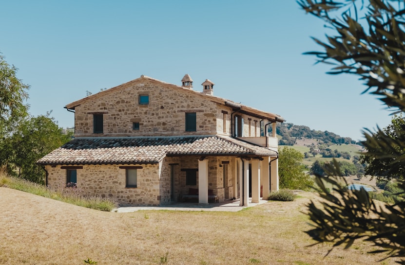 A rustic two-story stone house with a tile roof surrounded by a grassy landscape and distant hills. The structure features multiple windows, a porch with columns, and two small chimney-like structures on the roof. Trees are visible in the foreground and background.