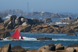 A small sailboat with vibrant red sails navigates through a rocky sea area. The background features rugged rock formations and distant lighthouses under a clear blue sky.