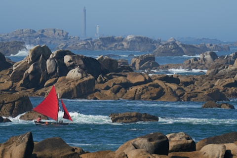 A small sailboat with vibrant red sails navigates through a rocky sea area. The background features rugged rock formations and distant lighthouses under a clear blue sky.
