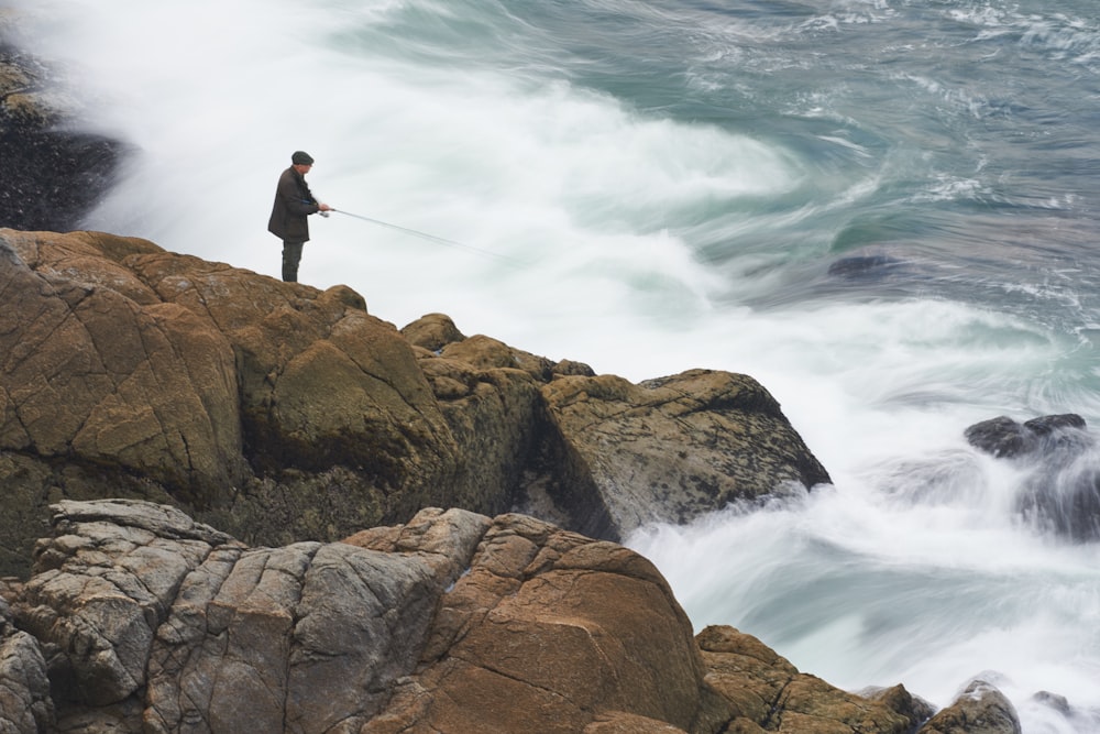 man in black jacket standing on brown rock formation near body of water during daytime