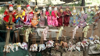A collection of colorful traditional puppets displayed on wooden racks. The puppets feature intricate costumes with vibrant colors such as red, yellow, pink, and green. They include ornate details like embroidered patterns and headdresses, some with wings. The lower section features marionette animals, predominantly elephants, in various playful poses.