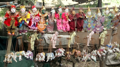 A collection of colorful traditional puppets displayed on wooden racks. The puppets feature intricate costumes with vibrant colors such as red, yellow, pink, and green. They include ornate details like embroidered patterns and headdresses, some with wings. The lower section features marionette animals, predominantly elephants, in various playful poses.