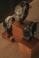 Three elegant watches are displayed on wooden blocks. Each watch features a metallic band and a round face, with varied designs and color schemes. The arrangement showcases the watches from different angles, emphasizing their craftsmanship and style.