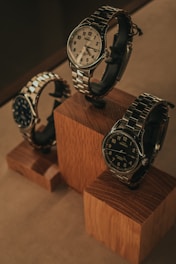 Three elegant watches are displayed on wooden blocks. Each watch features a metallic band and a round face, with varied designs and color schemes. The arrangement showcases the watches from different angles, emphasizing their craftsmanship and style.