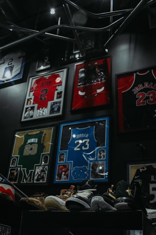 Framed sports jerseys, including a red one with the name and number Jeter 2, a red Bulls jersey with Jordan 23, a yellow and green jersey with James 23, and a blue jersey with Jordan 23. There are multiple sports memorabilia and photographs surrounding the jerseys. Several pairs of colorful athletic shoes are displayed on a stand at the bottom of the image.