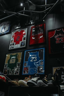 Framed sports jerseys, including a red one with the name and number Jeter 2, a red Bulls jersey with Jordan 23, a yellow and green jersey with James 23, and a blue jersey with Jordan 23. There are multiple sports memorabilia and photographs surrounding the jerseys. Several pairs of colorful athletic shoes are displayed on a stand at the bottom of the image.