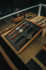 A collection of five wristwatches neatly arranged on a gray felt-lined wooden tray within a glass display case. The watches have various designs and styles, with brown and black leather straps. The lighting creates a warm and elegant ambiance, highlighting the watches and their details.
