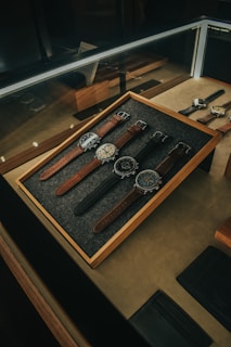 A collection of five wristwatches neatly arranged on a gray felt-lined wooden tray within a glass display case. The watches have various designs and styles, with brown and black leather straps. The lighting creates a warm and elegant ambiance, highlighting the watches and their details.