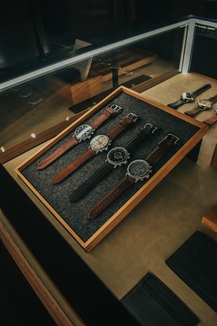A collection of five wristwatches neatly arranged on a gray felt-lined wooden tray within a glass display case. The watches have various designs and styles, with brown and black leather straps. The lighting creates a warm and elegant ambiance, highlighting the watches and their details.