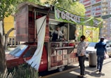 A street food vendor operates out of a red vehicle converted into a food truck, with a large green banner indicating the sale of 'tortas'. A customer stands at the counter waiting for service, and another person walks by carrying a bag. The scene is shaded by nearby trees and buildings in the background.