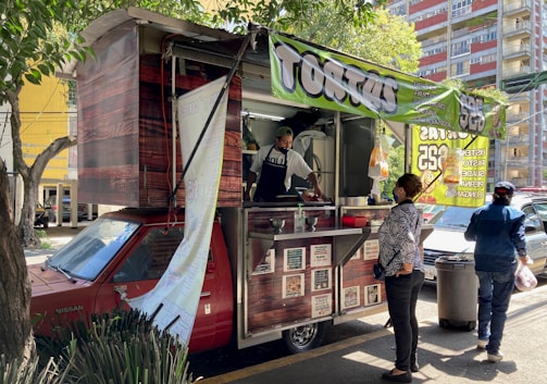 A street food vendor operates out of a red vehicle converted into a food truck, with a large green banner indicating the sale of 'tortas'. A customer stands at the counter waiting for service, and another person walks by carrying a bag. The scene is shaded by nearby trees and buildings in the background.