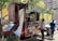 A street food vendor operates out of a red vehicle converted into a food truck, with a large green banner indicating the sale of 'tortas'. A customer stands at the counter waiting for service, and another person walks by carrying a bag. The scene is shaded by nearby trees and buildings in the background.