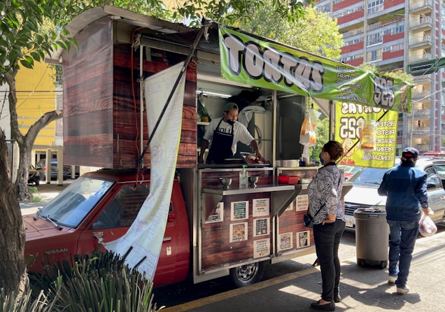 A street food vendor operates out of a red vehicle converted into a food truck, with a large green banner indicating the sale of 'tortas'. A customer stands at the counter waiting for service, and another person walks by carrying a bag. The scene is shaded by nearby trees and buildings in the background.