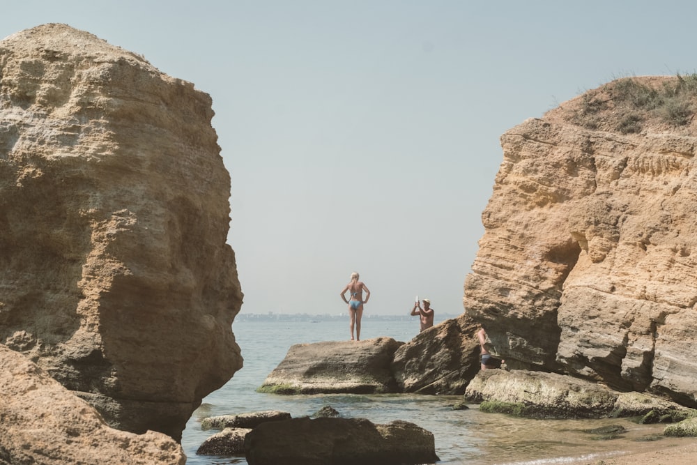 woman in black tank top and black shorts standing on brown rock formation near body of during