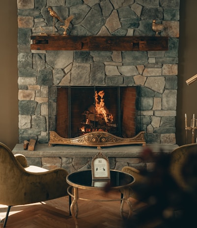 A cozy stone fireplace with a warm fire burning, surrounded by elegant decor. The mantel features decorative bird sculptures, and two plush chairs face a small round table with a framed picture or menu. The ambiance is inviting and luxurious, with soft lighting enhancing the stone textures.