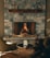 A cozy stone fireplace with a warm fire burning, surrounded by elegant decor. The mantel features decorative bird sculptures, and two plush chairs face a small round table with a framed picture or menu. The ambiance is inviting and luxurious, with soft lighting enhancing the stone textures.