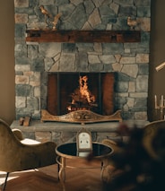 A cozy stone fireplace with a warm fire burning, surrounded by elegant decor. The mantel features decorative bird sculptures, and two plush chairs face a small round table with a framed picture or menu. The ambiance is inviting and luxurious, with soft lighting enhancing the stone textures.