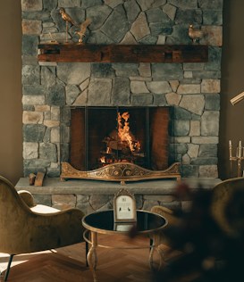 A cozy stone fireplace with a warm fire burning, surrounded by elegant decor. The mantel features decorative bird sculptures, and two plush chairs face a small round table with a framed picture or menu. The ambiance is inviting and luxurious, with soft lighting enhancing the stone textures.