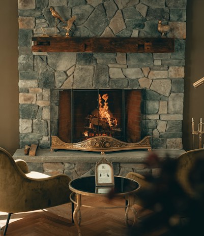 A cozy stone fireplace with a warm fire burning, surrounded by elegant decor. The mantel features decorative bird sculptures, and two plush chairs face a small round table with a framed picture or menu. The ambiance is inviting and luxurious, with soft lighting enhancing the stone textures.