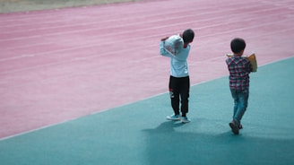 Two children walk along a track, one wearing a white shirt and black pants carrying a large bag, the other in a plaid shirt holding a book or folder. The background features a pink running track, while they walk on a green surface.