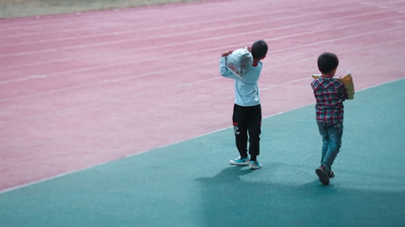 Two children walk along a track, one wearing a white shirt and black pants carrying a large bag, the other in a plaid shirt holding a book or folder. The background features a pink running track, while they walk on a green surface.