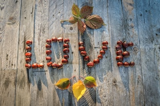 The word 'LOVE' is spelled out using chestnuts on a wooden surface. Above and below the letters are autumn leaves in various colors, including shades of red and yellow. The wooden planks are weathered and textured, adding a rustic feel to the arrangement.