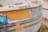 A close-up view of an old, weathered boat with peeling paint, revealing layers of blue, orange, and white colors. The surface shows signs of aging and exposure to the elements, with rust spots and scratches visible.