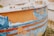 A close-up view of an old, weathered boat with peeling paint, revealing layers of blue, orange, and white colors. The surface shows signs of aging and exposure to the elements, with rust spots and scratches visible.