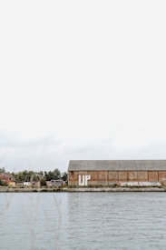 A large, rustic warehouse with graffiti on its walls stands next to a calm body of water. The word 'UP' is prominently painted on the building. Trees and some industrial equipment are visible in the background under a cloudy sky.