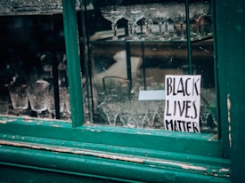 A storefront window displays various crystal glasses arranged neatly inside. A handwritten sign with the text 'Black Lives Matter' is taped to the glass. The window frame is painted in a worn green color.