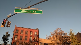 A street scene with a green street sign displaying 'Atlantic Av' hanging from a traffic light pole. Below the sign is a red-brick building with multiple windows and an external fire escape, surrounded by trees. The sky is clear and illuminated by the warm glow of sunset, casting a golden hue over the scene.