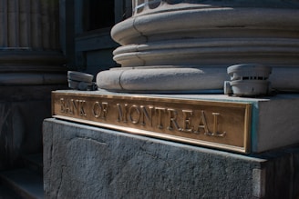 An exterior view of the Bank of Montreal, featuring a decorative stone column and a bronze plaque with engraved text. Two security cameras are positioned on either side of the plaque.