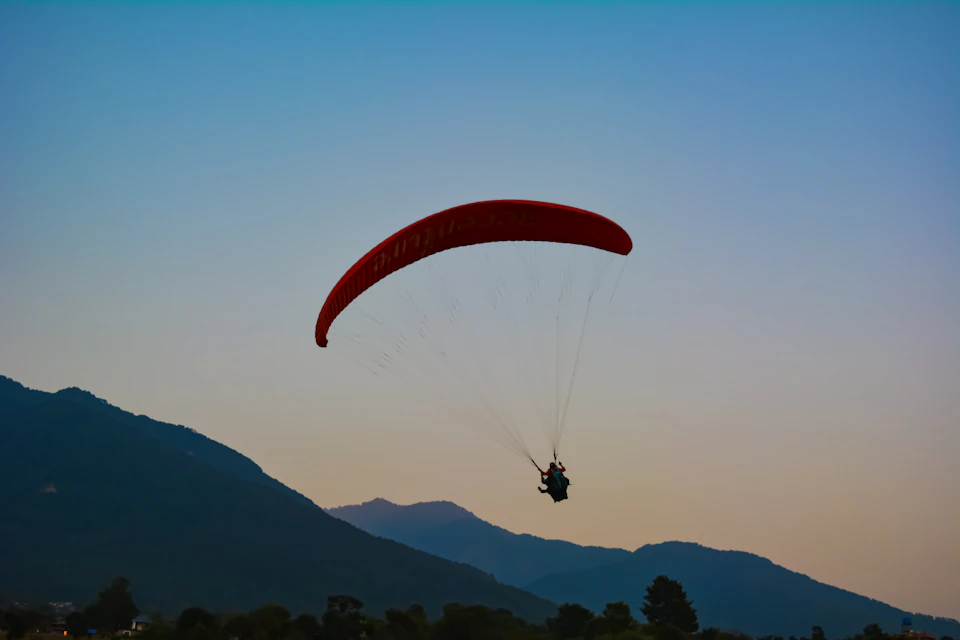 Red parachute soars through the sky with two people inside above a mountainous landscape