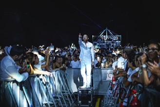 A man dressed in white stands on a platform among a crowd at a concert or event. He appears to be performing or addressing the audience, with one hand pointing forward. The crowd is gathered on both sides of a barrier, with many people filming or taking photos using their phones. The scene is illuminated with stage lighting, enhancing the vibrant atmosphere.