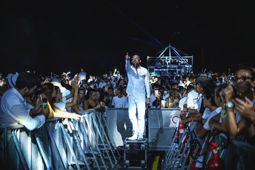 A man dressed in white stands on a platform among a crowd at a concert or event. He appears to be performing or addressing the audience, with one hand pointing forward. The crowd is gathered on both sides of a barrier, with many people filming or taking photos using their phones. The scene is illuminated with stage lighting, enhancing the vibrant atmosphere.
