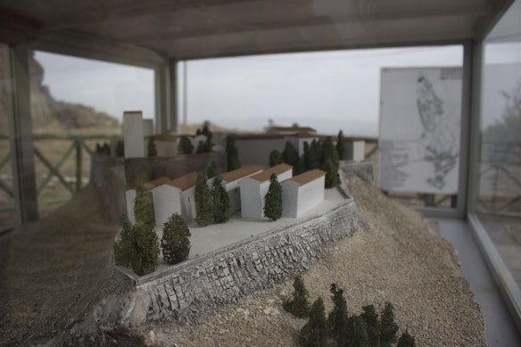 A detailed miniature architectural model depicts a hillside terrain with various structures, possibly houses or buildings, surrounded by miniature trees. The model is enclosed in a glass case with a railing visible in the background. A map or information board is located on the right side outside the glass case.